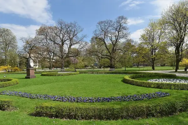 Piękny park miejski z rzeźbą, kwitnącymi rabatami i zielonymi trawnikami.