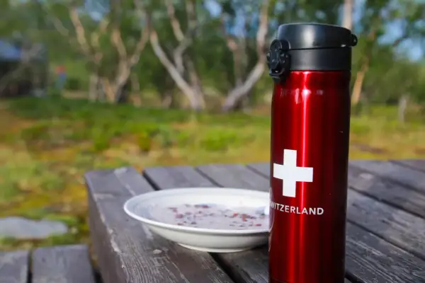 Röd termos med schweizisk flagga och en skål gröt på ett träbord. Perfekt för en frukost vandring i naturen.