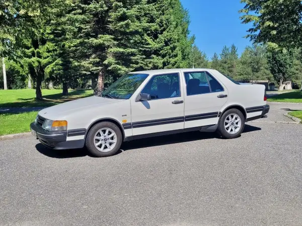 Vit Saab 9000 CD parkerad på en asfalterad väg med gröna träd och en blå himmel i bakgrunden.