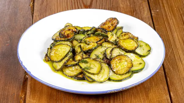 Piatto di zucchine alla poverella, fette sottili e dorate condite con olio e erbe aromatiche, su un piatto bianco con bordo blu.