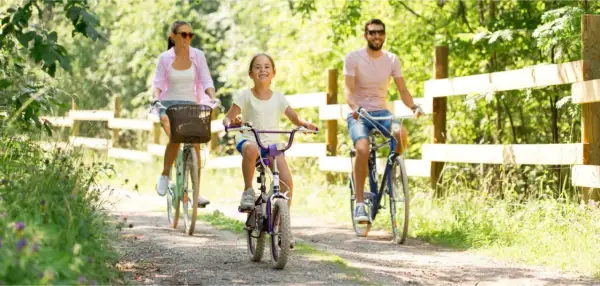 Rodzina cieszy się rodzinnym wyjazdem na **Mazury rowerem**. Radosna dziewczynka jedzie na fioletowym rowerze, a jej rodzice podążają za nią.