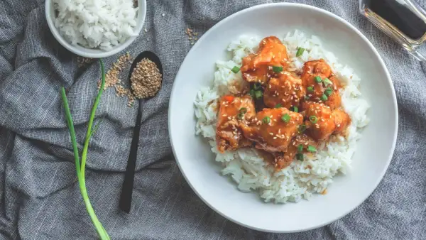 Ein Teller mit Reis und Hähnchen in süß-saurer Sauce, bestreut mit Sesam und Frühlingszwiebeln. Dieses Gericht hat Reis, Proteine und ist ein Genuss.