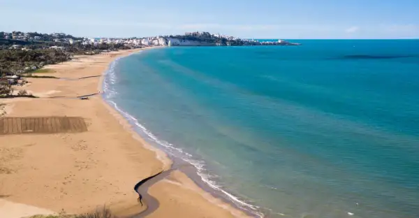 La spiaggia della Scialara si estende con sabbia dorata e mare azzurro, con il borgo bianco arroccato sulla scogliera in lontananza.