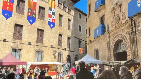Gente disfruta del mercado medieval de las tres culturas, con puestos y banderas históricas adornando edificios antiguos.