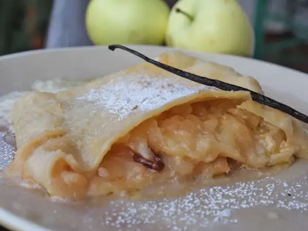 Ein Stück bayerischer Apfelstrudel, bestäubt mit Puderzucker und garniert mit einer Vanilleschote, daneben zwei Äpfel.