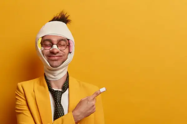 Uomo con benda in testa e cerotto sul dito indica come viene pagato l'infortunio, con un sorriso ironico.