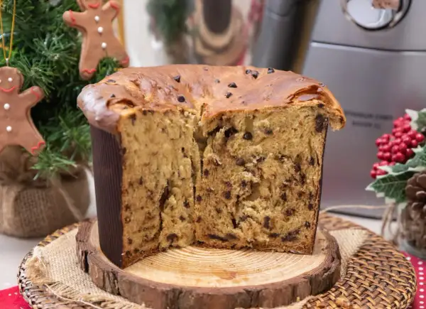 Un soffice panettone natalizio con gocce di cioccolato, tagliato a metà, su un tagliere di legno. Decorazioni natalizie sullo sfondo.