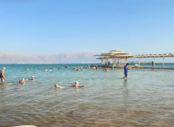 Ludzie unoszą się na wodzie Morza Martwego, korzystając z jego niezwykłego zasolenia. W tle góry i plaża z zacienionymi miejscami.