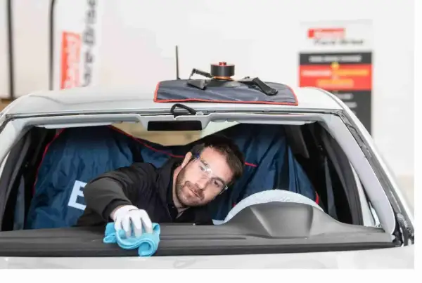Un technicien porte des lunettes de protection et utilise un chiffon bleu pour nettoyer l'intérieur du pare-brise d'une voiture.