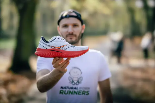 Mężczyzna w koszulce "Natural Born Runners" prezentuje biało-czerwony but biegowy Saucony. Saucony to znana firma z tradycjami.