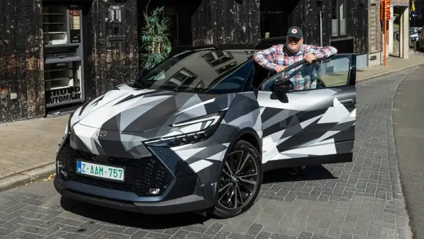 Man lutar sig mot en Toyota C-HR Hybrid i kamouflagemönster. Bilen är parkerad på en kullerstensgata framför en byggnad med en automat.