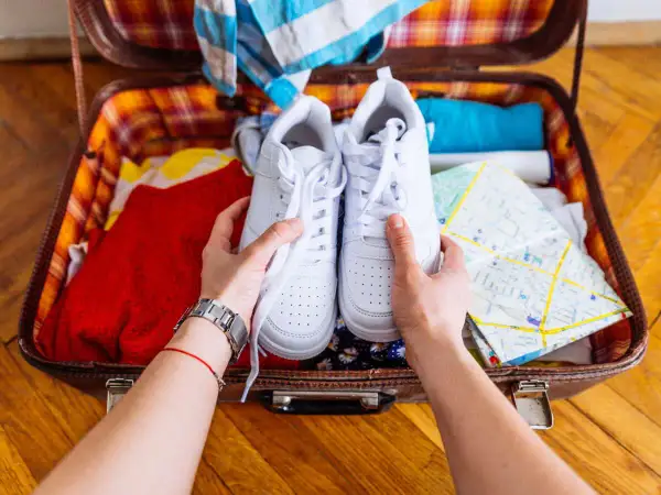Comment ranger des chaussures dans une valise : une paire de baskets blanches est tenue au-dessus d'une valise ouverte remplie de vêtements et d'une carte.