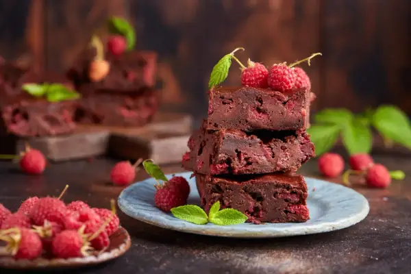 Czekoladowe brownie z malinami, ułożone na talerzyku, ozdobione świeżymi malinami i listkami mięty.