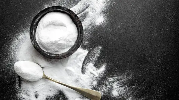 Biały proszek, prawdopodobnie soda oczyszczona na mrówki, w miseczce i na łyżce na czarnym tle.