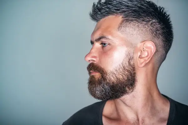 Mann mit Bart und tiefer fasson haarschnitt, im Profil auf grauem Hintergrund.