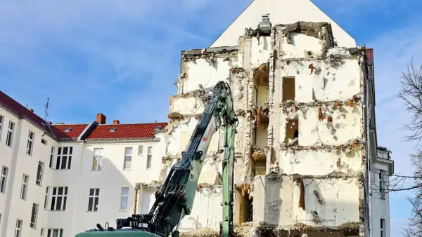 Koparka pracuje przy rozbiórce budynku, odsłaniając jego zniszczoną konstrukcję.