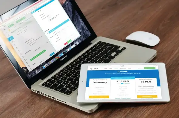 Programista .NET pracuje nad aplikacją na laptopie i tablecie. Na ekranach widoczne są formularze i cennik usług.