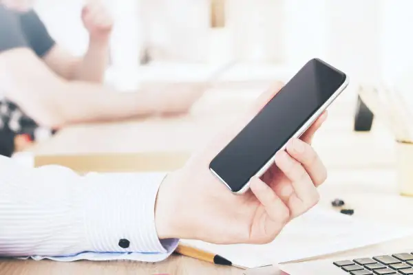 Dłoń trzyma smartfon, być może przeglądając ofertę kredyt orange. W tle widać biurko i inne osoby.