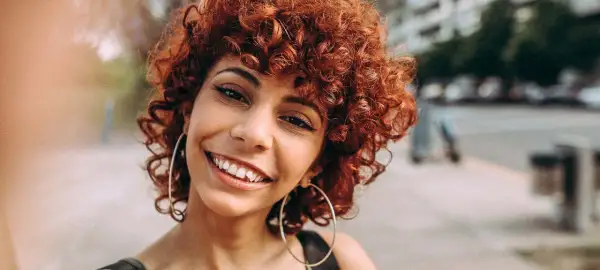 Sonrisa radiante de una mujer con rizos castaño rojizo y grandes aros plateados.
