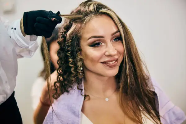 Fryzjerka tworzy loki na włosach z **chunky highlights**. Uśmiechnięta kobieta z makijażem i kolczykiem w wardze.