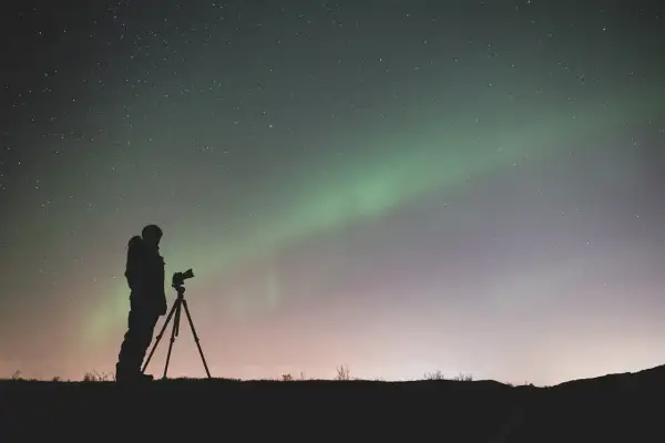 Sylwetka fotografa z aparatem na statywie pod rozgwieżdżonym niebem i zorzą polarną. Efekt bokeh gwiazd dodaje magii.