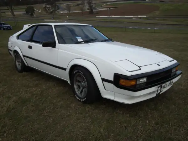 Biała Toyota Supra MK2 z lat 80. stoi na trawie, z widokiem na tor wyścigowy w tle.
