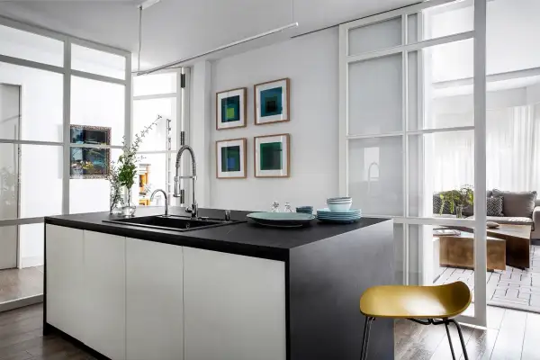 Isla de cocina moderna con encimera oscura, separada del salón por una pared de cristal.