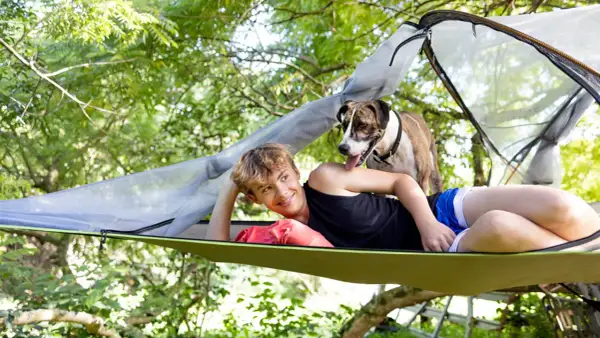 Un chico sonríe mientras su perro se asoma por encima de él en una hamaca con mosquitera, disfrutando del aire libre.