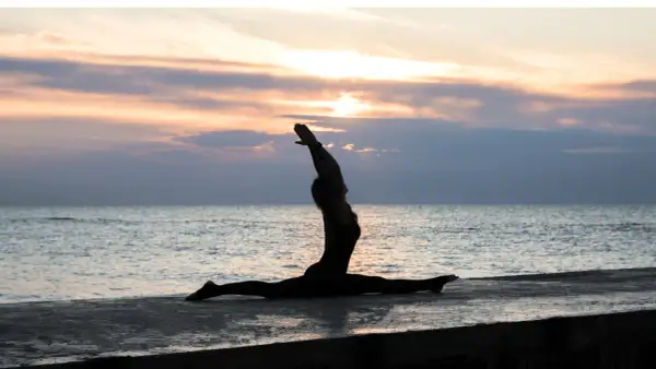 Silhouette di una donna in Ardha Hanumanasana sulla riva del mare al tramonto, con le braccia alzate verso il cielo.