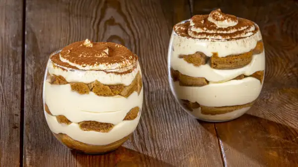 Due bicchieri di tiramisù senza uova, stratificati con crema e savoiardi, spolverati di cacao, su un tavolo di legno.