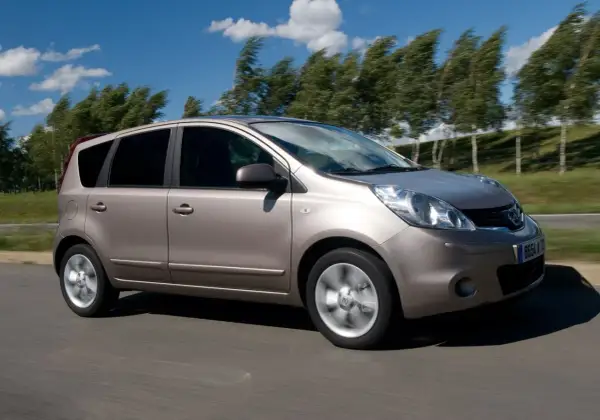 Złoty Nissan Note 2010 pędzi po drodze, drzewa na tle falują na wietrze.