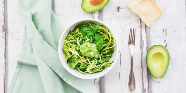 Zoodles con pesto di avocado, guarniti con pinoli e basilico. Accanto, mezzo avocado, un pezzo di parmigiano e una forchetta.
