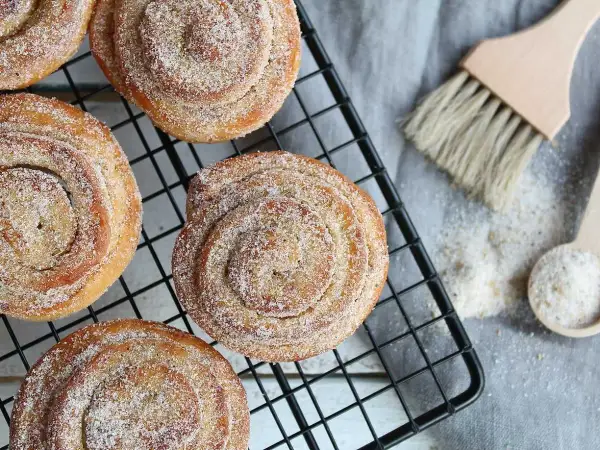 Nybakade kanelbullar med strössel av brunt socker och pärlsocker på ett svart galler. En träpensel och en sked med socker ligger bredvid.