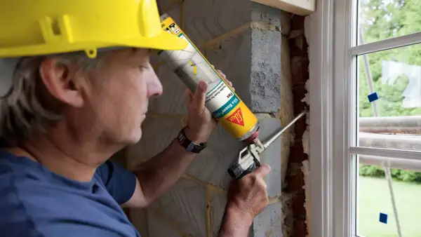 Hombre con casco amarillo usa sellador de poliuretano para rellenar huecos en instalación de ventana.