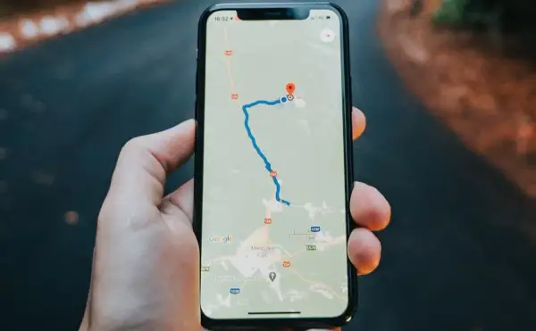 Dłoń trzyma iPhone'a z mapą Google, pokazującą trasę i lokalizację.