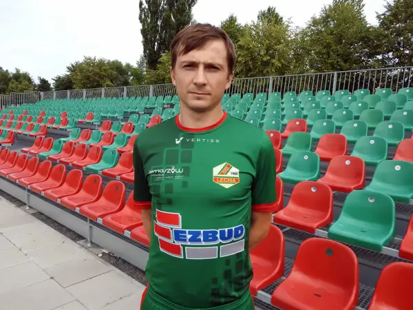 Kamil Szymczak w zielonej koszulce sportowej stoi przed rzędem czerwonych i zielonych krzesełek na stadionie.