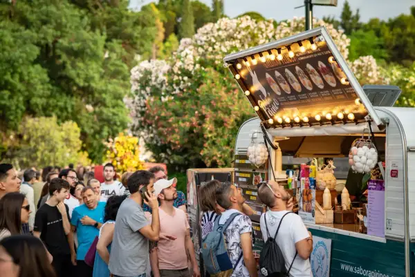 Gente disfrutando de food trucks en Zaragoza, un evento donde la gastronomía florece entre árboles y flores.