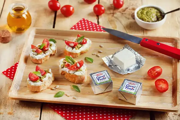 Bruschettas garnies de fromage frais, tomates et pesto, avec des emballages de Carré Frais le cendre et des tomates cerises sur un plateau en bois.