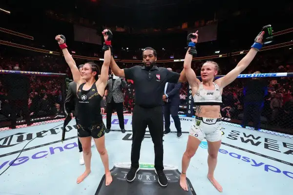 Two female fighters stand with arms raised, a referee between them. The outcome of their boxing draw is announced to the roaring crowd.