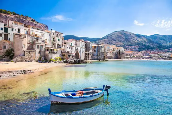 La **spiaggia più bella** di Cefalù, con case colorate a picco sul mare, una barca bianca e blu e acque cristalline.