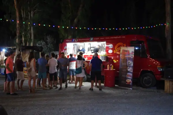 Gente disfrutando en un food trucks festival bajo luces de colores. Un camión rojo sirve comida mientras clientes esperan.