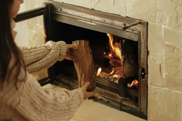 Une personne en pull ajoute une bûCHE dans un poêle à bois qui fume, porte ouverte. La chaleur des flammes réchauffe la pièce.