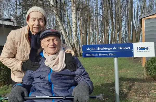 Uśmiechnięta para seniorów pozuje przy tablicy "Alejka Hrabiego de Rosset" w dom seniora Rotmanka.