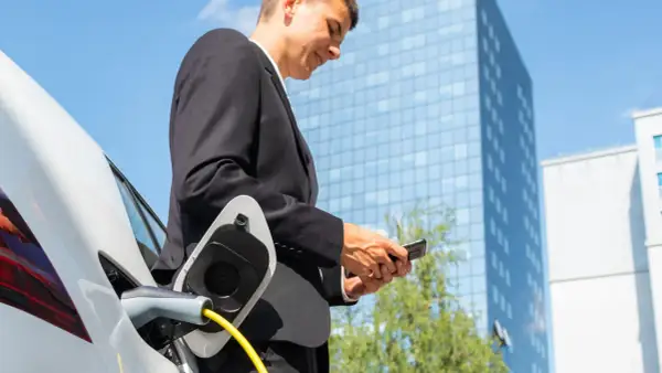 Uomo in giacca e cravatta controlla il telefono mentre la sua auto elettrica si ricarica, pianificare viaggio auto elettrica è facile con la tecnologia.