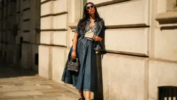 Mujer con gafas de sol posa apoyada en una pared, luciendo un conjunto vaquero y una falda vaquera larga.