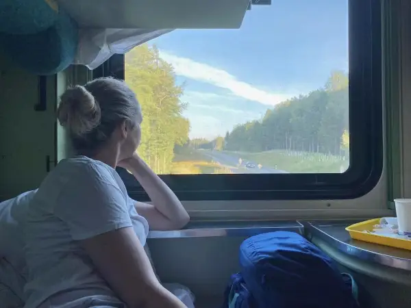 Frau blickt aus dem Fenster eines Nachtzuges nach Italien. Die Landschaft zieht vorbei, Bäume und eine Straße sind zu sehen.