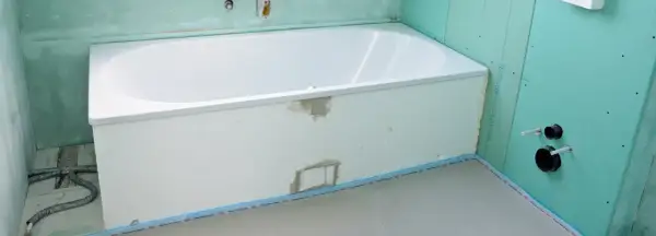 Einbau einer neuen, weißen Badewanne in einem Badezimmer im Rohbau. Grüne Gipskartonplatten und vorbereitete Rohre sind sichtbar.