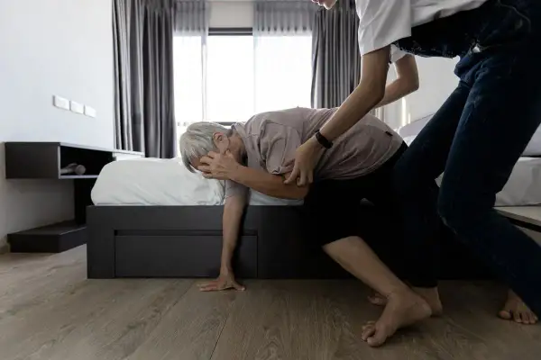 Assistenza a un anziano che accusa un cedimento delle gambe, con una persona che lo sorregge vicino al letto.