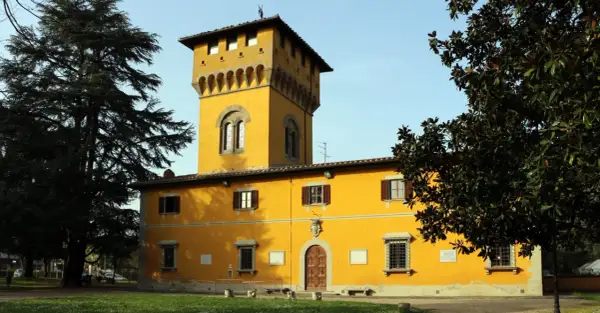 Edificio giallo con torre merlata a Borgo San Lorenzo, circondato da alberi verdi e un cielo azzurro.