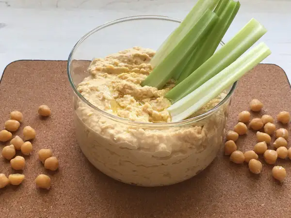 Pyszny, domowy hummus przepis. Kremowa pasta z ciecierzycy z łodygami selera i ziarnami na korkowej macie.